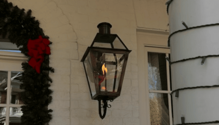 Lit black gas lantern mounted on a white brick wall next to a Christmas wreath with a red bow and a column wrapped in string lights.