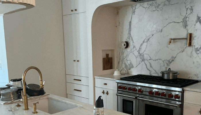 Modern white kitchen with marble backsplash, brass faucet, pot filler, and stainless steel range with red knobs.